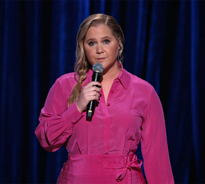 Amy Schumer on stage wearing pink, speaking into a microphone while showcasing new weight loss jab results. Amy Schumer on stage wearing pink, speaking into a microphone while showcasing new weight loss jab results.