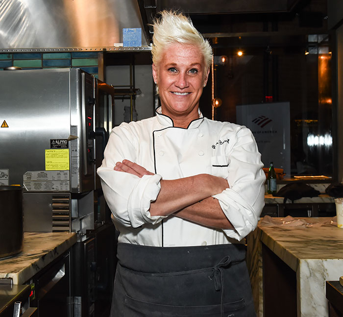 Master Chef Anne Burrell in kitchen wearing white chef coat, smiling with arms crossed in a professional setting.