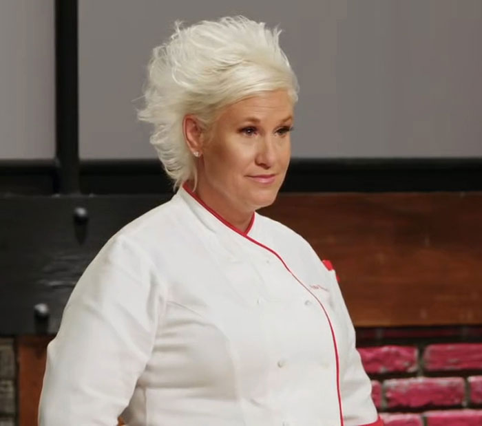 Master Chef Anne Burrell in white chef coat, looking thoughtful in kitchen setting related to medical examiner's report. - 2