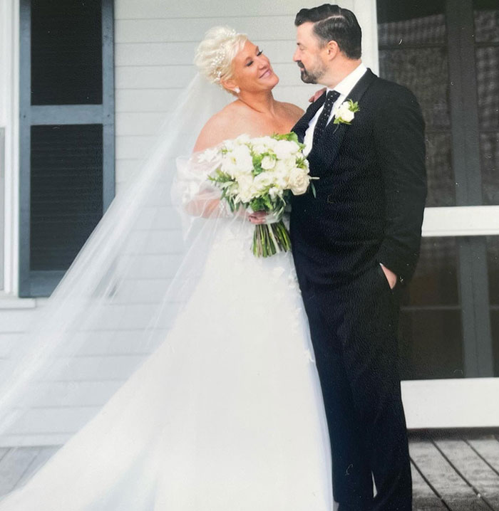 Bride in white gown holding bouquet looks at groom in black suit standing on porch, related to medical examiner's report. - 6