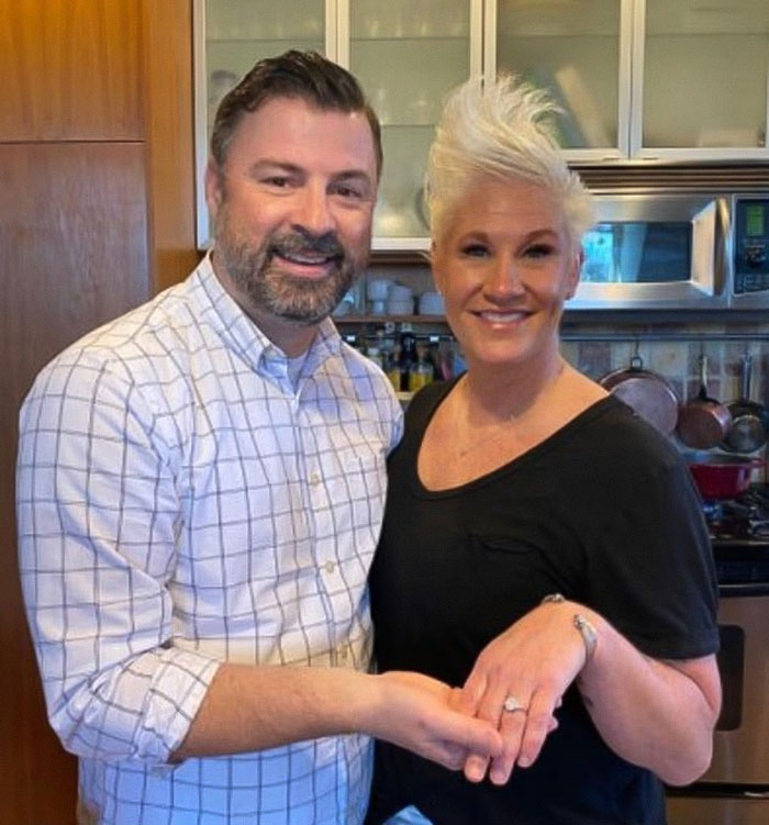 Master Chef Anne Burrell smiling in a kitchen with a man, both showing a ring on her finger. - 7