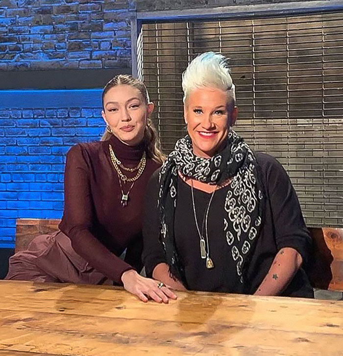 Master chef Anne Burrell with a guest, smiling and seated at a wooden table in a studio setting. - 10