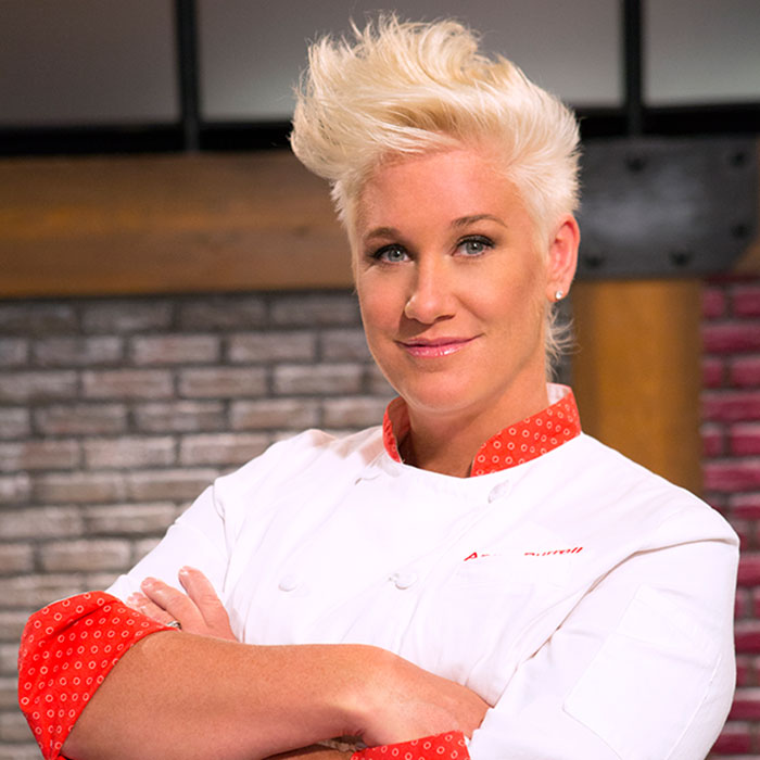 Master Chef Anne Burrell posing confidently in a white chef coat with a brick wall background. - 12