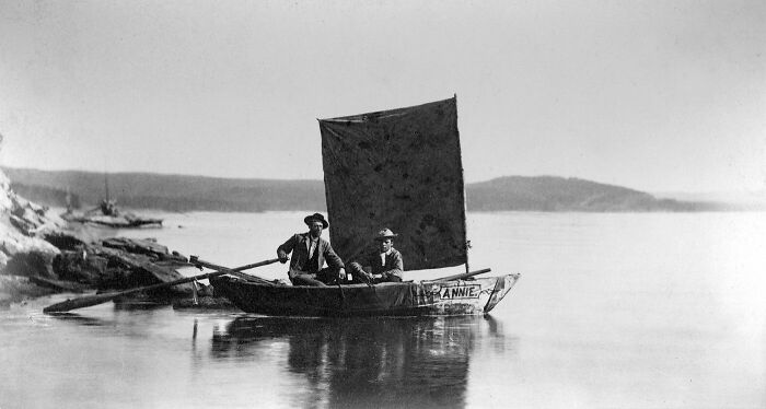 Two men sailing a small boat with a square sail on calm water in 1800s America historical scene