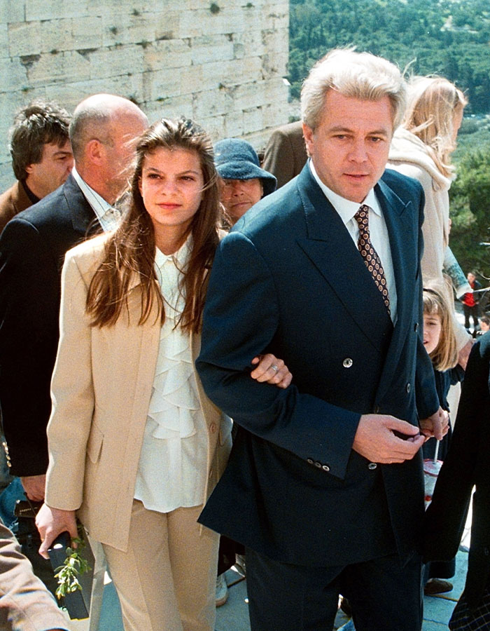 Athina Onassis, reclusive billionaire, pictured in a beige suit walking arm in arm during a rare public appearance.