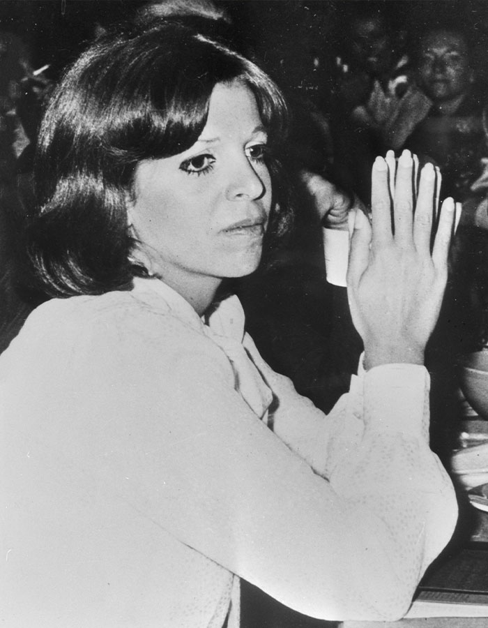 Reclusive billionaire Athina Onassis seated, wearing a light blouse, with hands raised in a reflective moment at an event.