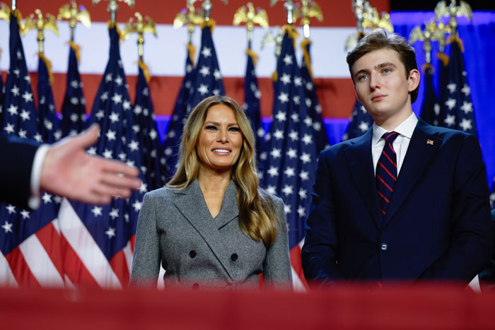 Barron Trump standing with Melania Trump in front of American flags highlighting luxury-filled childhood and public attention.