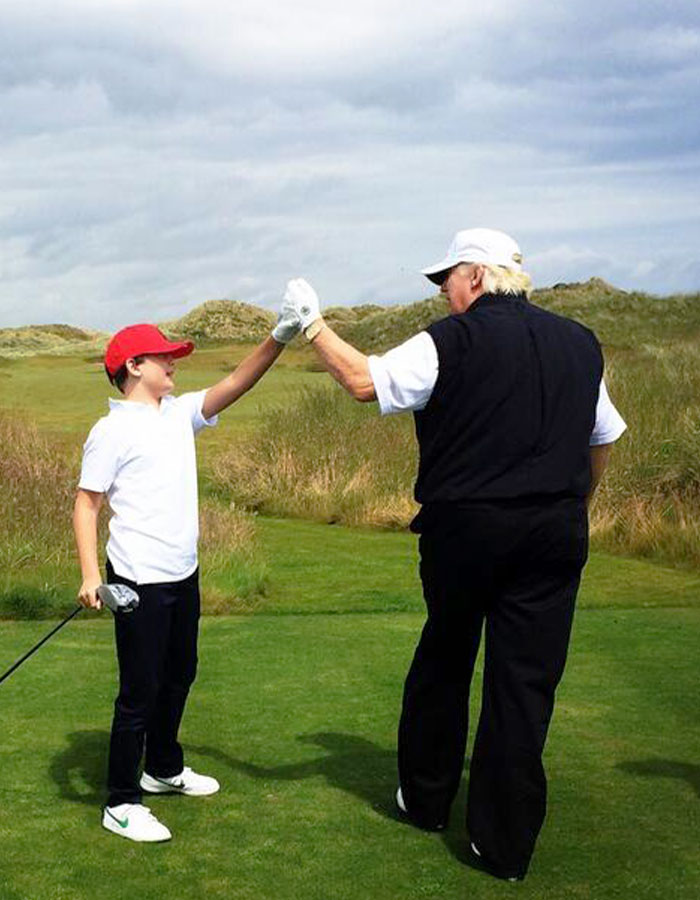 Barron Trump and an older man fist bump on a golf course, showcasing Barron's luxury-filled childhood outdoors.