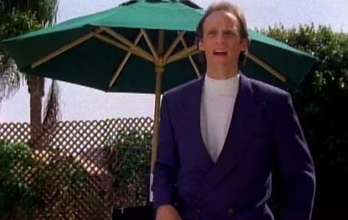 Man in a dark blazer standing outdoors under a green umbrella, with greenery and lattice fence in the background.