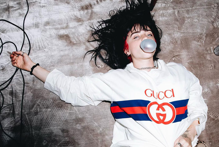 Billie Eilish lying down wearing a Gucci sweatshirt and blowing a bubble with gum, sparking nepo baby fan discussions.