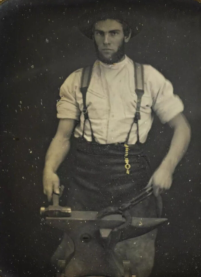 19th century photo of a blacksmith holding tongs and hammer, showcasing historical craftsmanship and period attire.