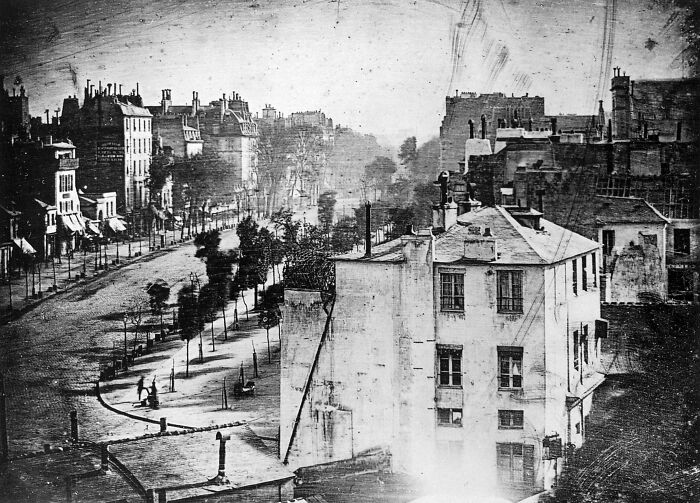 Early 19th century black and white photo showing a historic city street with old buildings and sparse trees lining the sidewalk.
