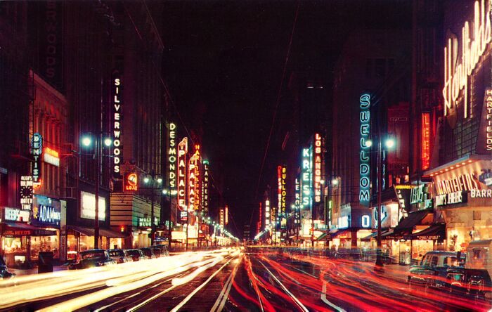 Nighttime city street scene in the 50s with vibrant neon signs and light trails showing daily life across the globe.