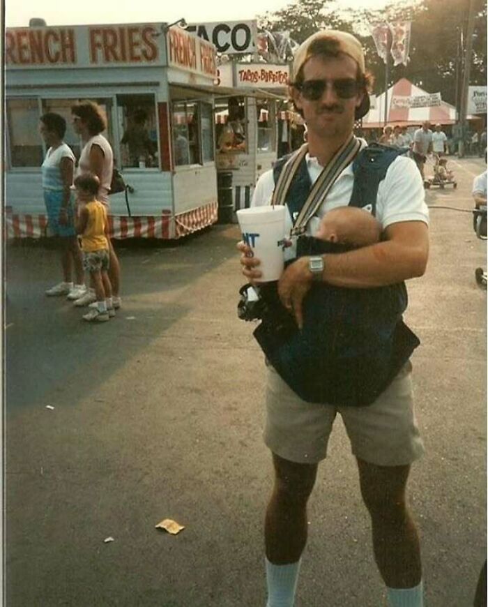 Man embodying old school cool dad style, wearing sunglasses and carrying baby in front carrier at outdoor fair.