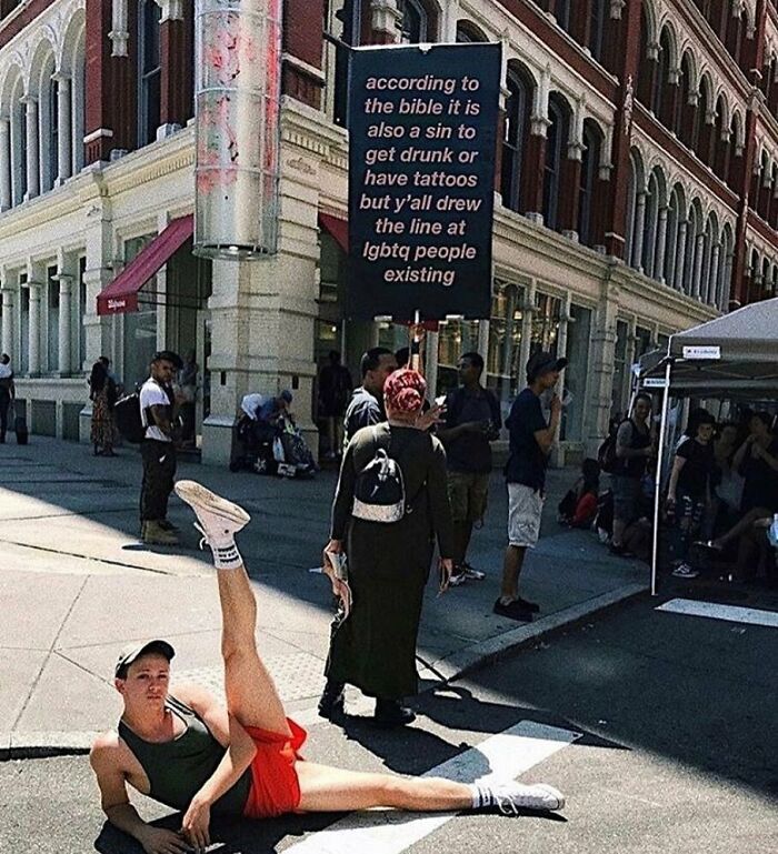 Person in athletic wear posing on the ground at a street protest with a sign about LGBTQ+ people in an urban setting.