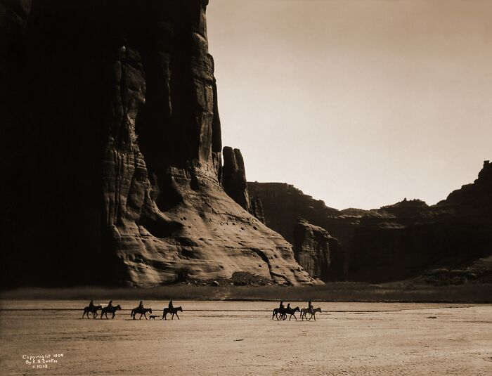 Vintage Native American family riding horses across a desert landscape with large rock formations in the background.