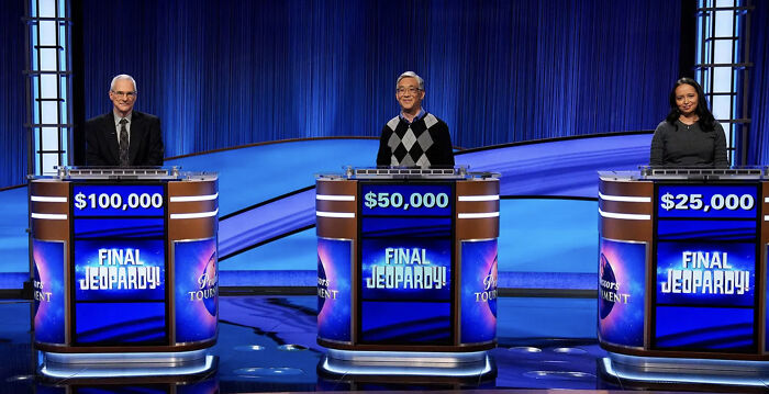 Three Jeopardy contestants standing behind podiums with Final Jeopardy screens showing their scores on the quiz show set.
