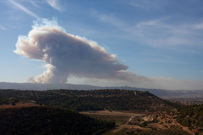 Smoke rising from a catastrophic wildfire over a dry landscape with hills and clear blue sky in the distance.
