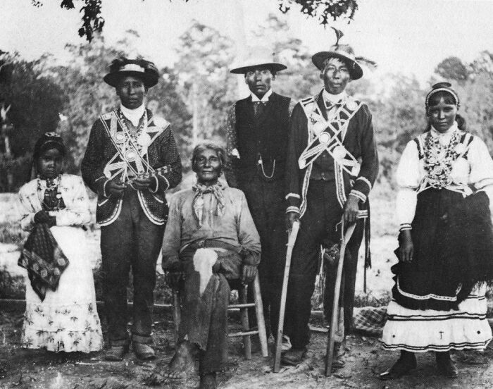 Vintage Native American family dressed in traditional clothing posing outdoors for a historical black and white photo.