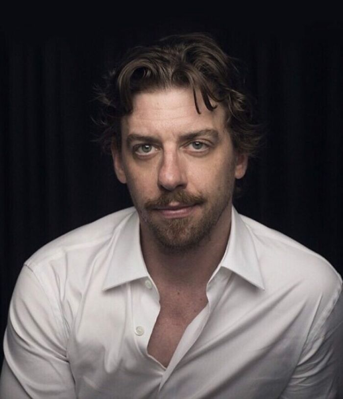 Man with tousled hair and beard wearing a white shirt, looking directly at the camera with a neutral expression.