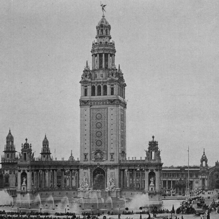 Historic old architecture with detailed tower and ornate sculptures in a grand public square filled with visitors.