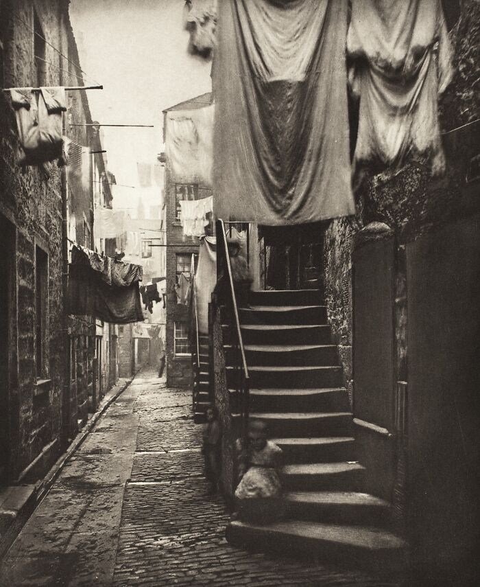 19th century photo of a narrow cobblestone street with hanging laundry and children sitting on stone stairs.