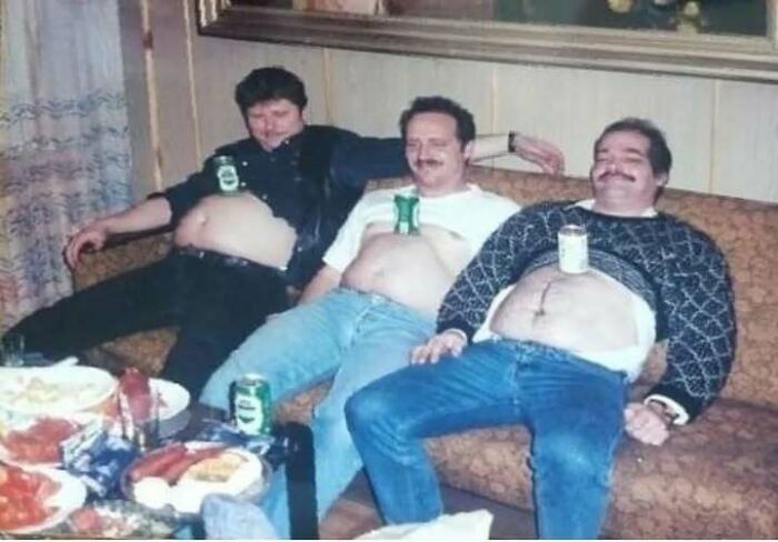 Three dads from the past showing old school cool vibes, sitting on a couch with drinks balancing on their stomachs.