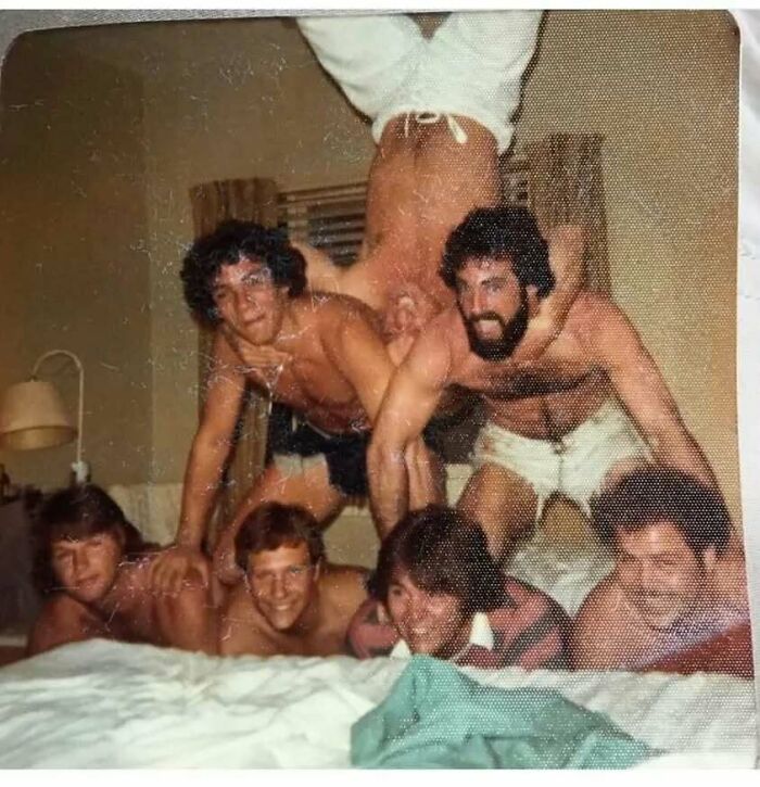 Group of old school cool dads from the past posing shirtless in a playful pyramid inside a vintage room.