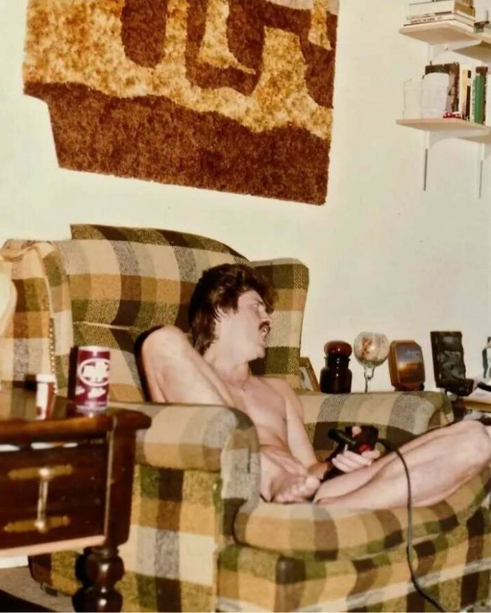 Shirtless dad from the past relaxing in a checkered chair playing video games in a vintage living room setting old school cool.