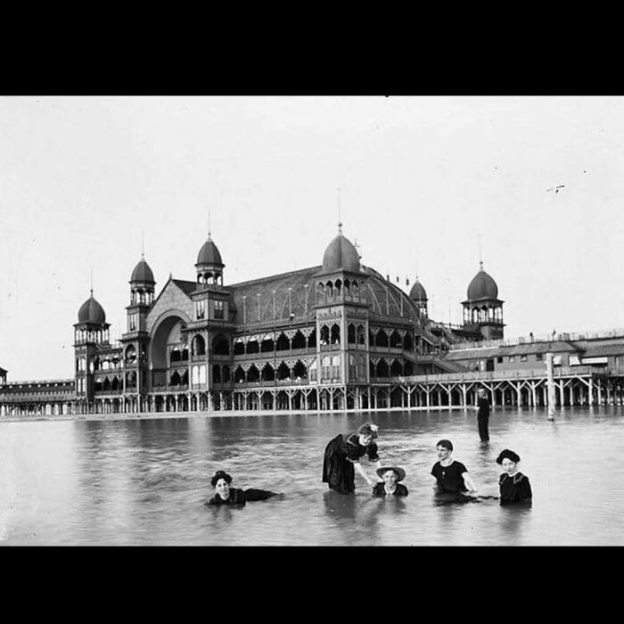 Black and white photo of old architecture with ornate domes and people bathing in water in front of the historic building.