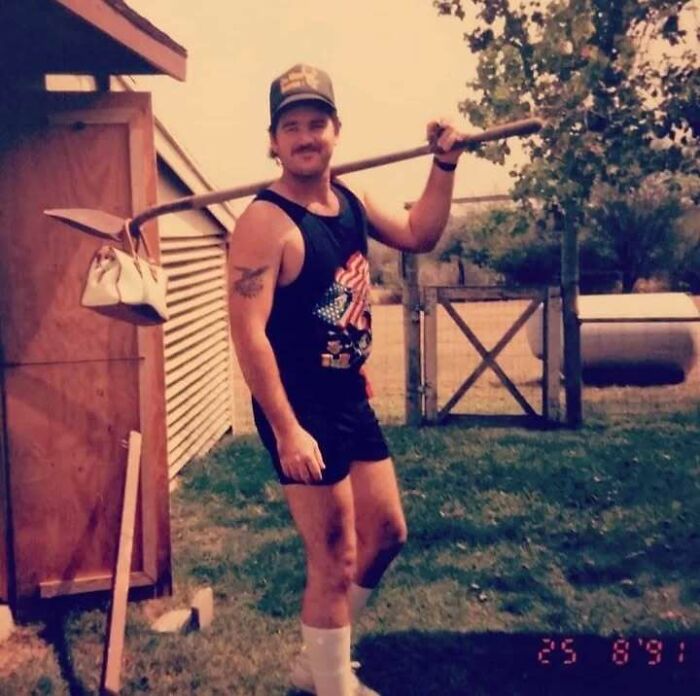 Man with mustache and tattoo wearing tank top and shorts, holding shovel over shoulder in backyard, showcasing old school cool dad style.