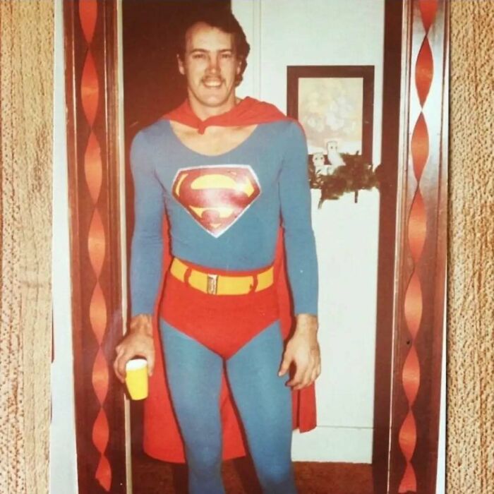 Man dressed in old school cool Superman costume from the past holding a drink and smiling indoors.