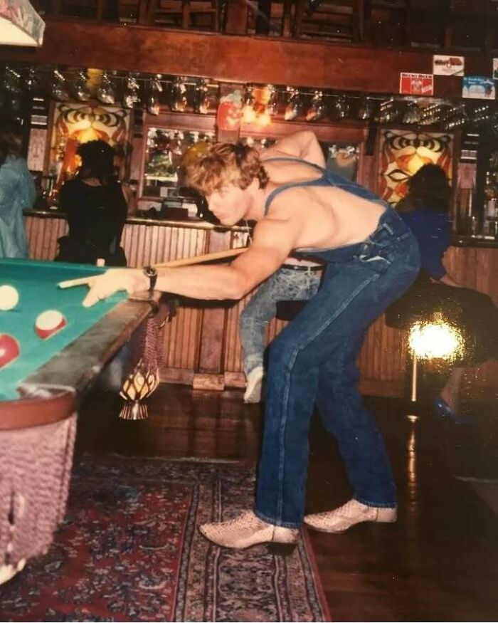 A shirtless dad in blue jeans and snakeskin boots playing pool in a vintage bar, capturing old school cool vibes.