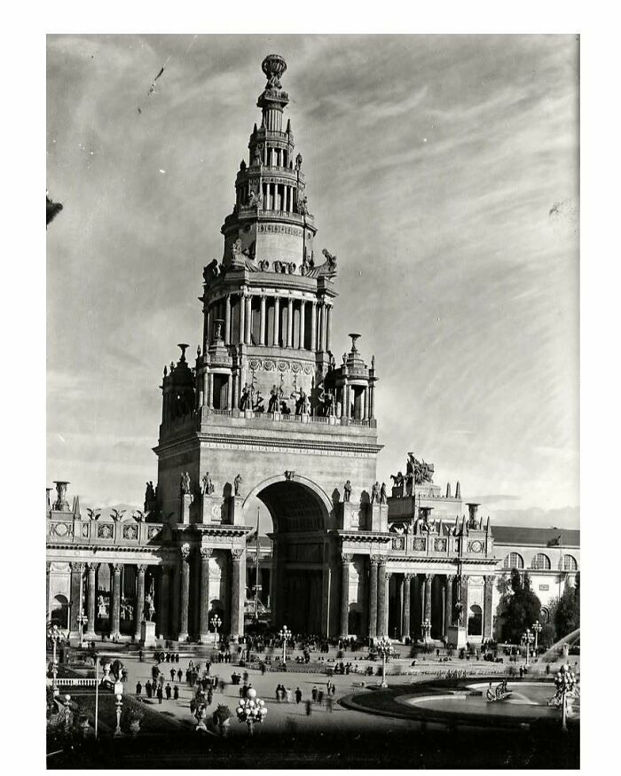 Black and white photo of old architecture featuring a grand ornate tower and arch with people gathered below.