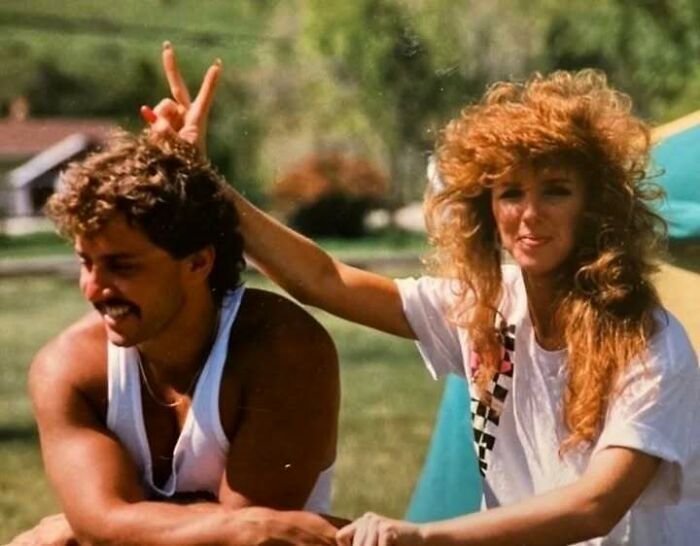 Vintage photo of a dad showing old school cool with mustache and sleeveless shirt outdoors with a woman making a playful gesture.