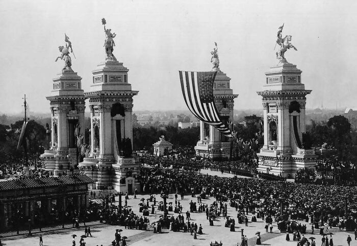 Crowd gathered around old architecture monuments with large statues and an American flag in a historic black and white photo.