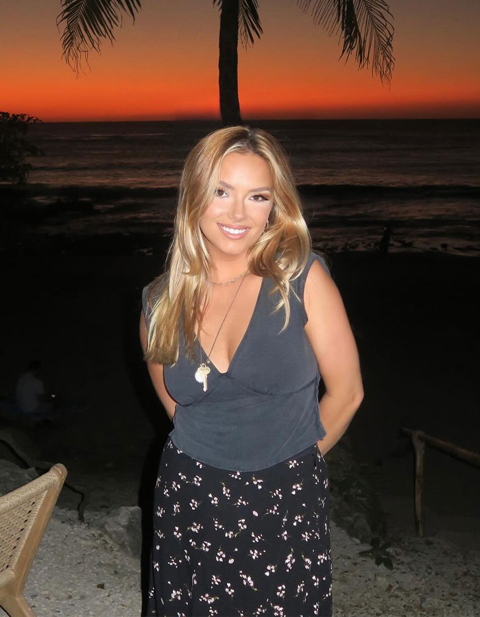 Young woman smiling outdoors at sunset with palm trees, related to Coldplay fan who exposed cheating CEO by filming kiss cam.