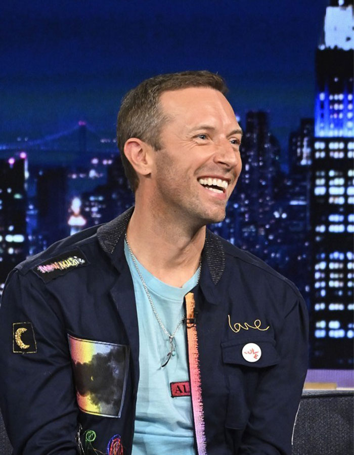 Chris Martin smiling in a dark jacket during a televised event with city lights blurred in the background.