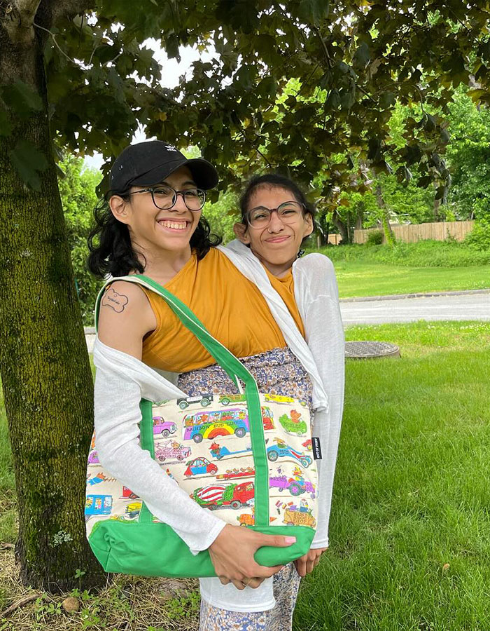 Conjoined twin influencer smiling outdoors wearing glasses and colorful outfit on a sunny day near a tree.