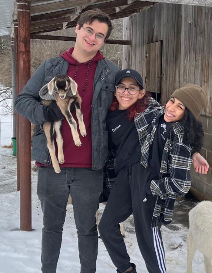 Conjoined twin influencer smiling outdoors with boyfriend and a small goat during a casual winter day.