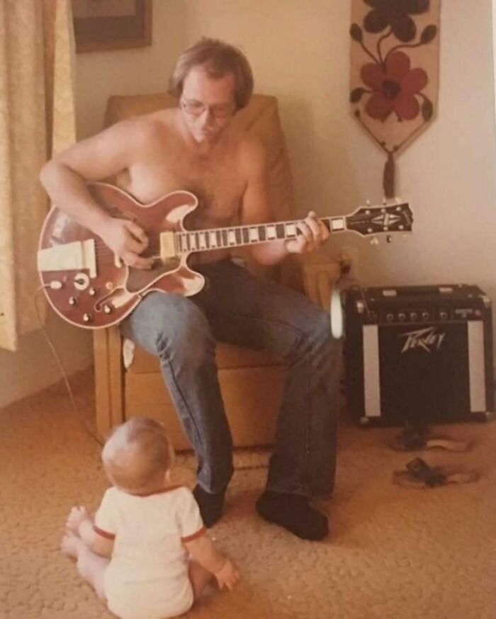 Shirtless dad playing electric guitar for baby on carpet, embodying old school cool with vintage amplifier nearby.