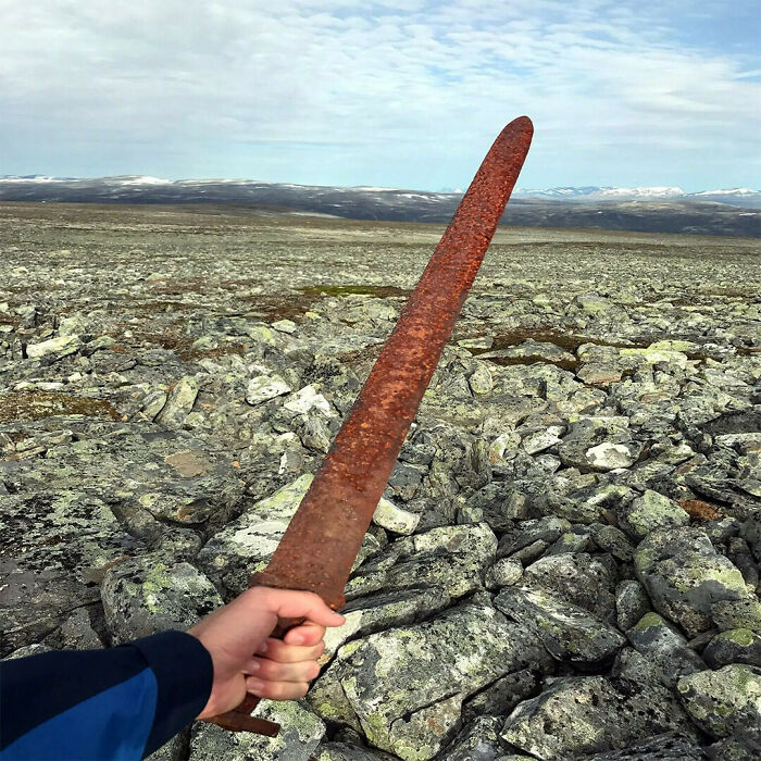 Hand holding a rusted iron sword over rocky terrain with mountains in the background, showcasing interesting facts.