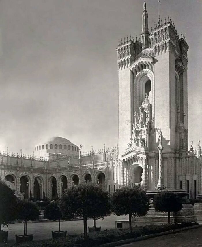Black and white photo of old architecture featuring detailed gothic tower and arched colonnade with manicured trees.