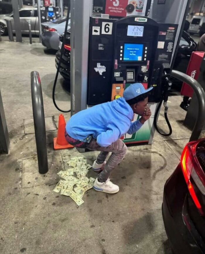 Young man squatting at gas station with dollar bills scattered beneath him, creating a confusing and unusual scene.
