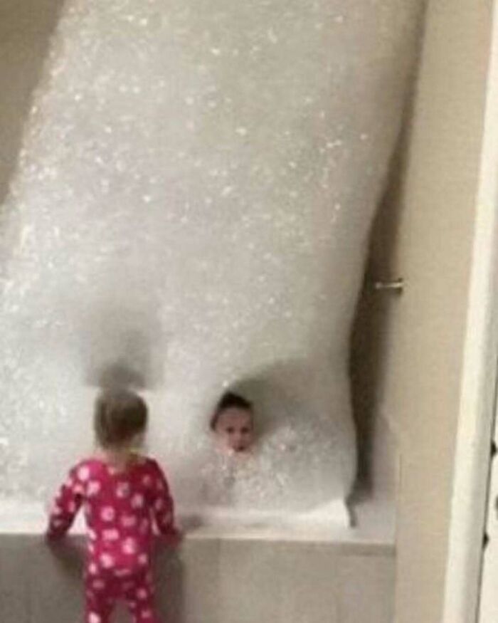 Child submerged in a bathtub full of bubbles with another child in pajamas looking on in a bizarre and funny scene.
