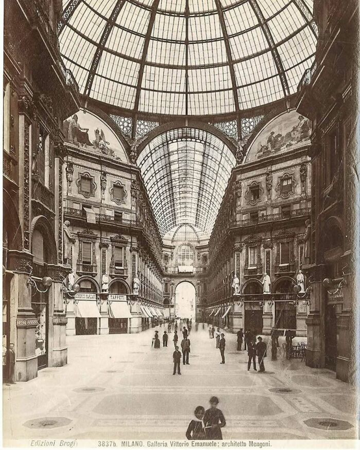 Historic old architecture inside a grand glass-domed gallery with ornate facades and people walking through.