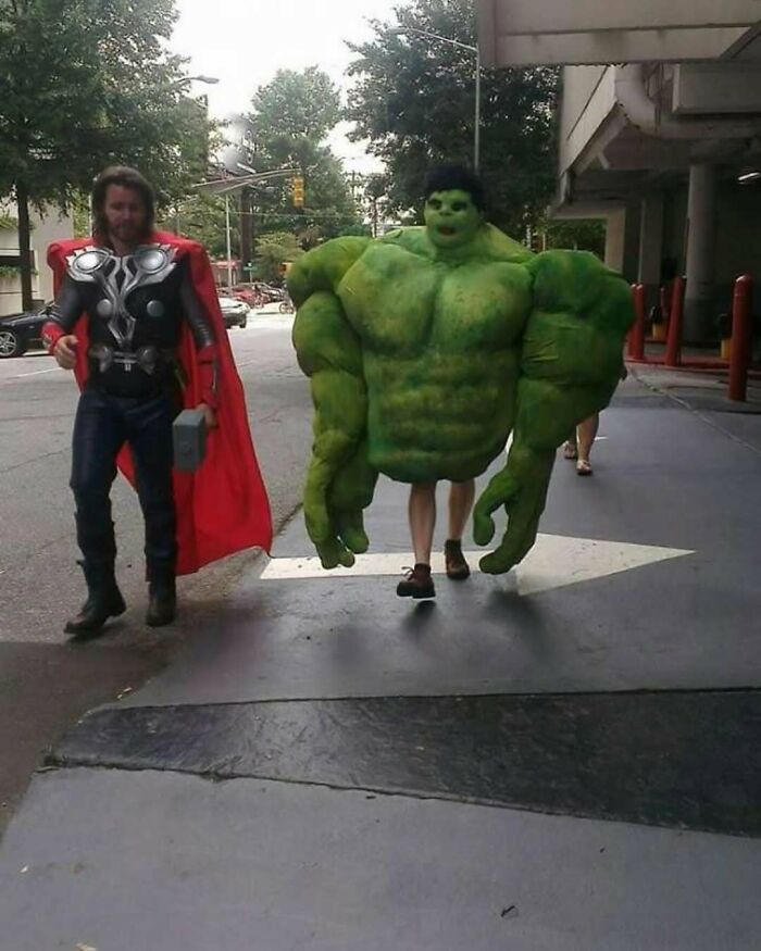 Two people dressed as oversized Hulk and Thor characters walking on a sidewalk in a bizarre scene from strange pics.