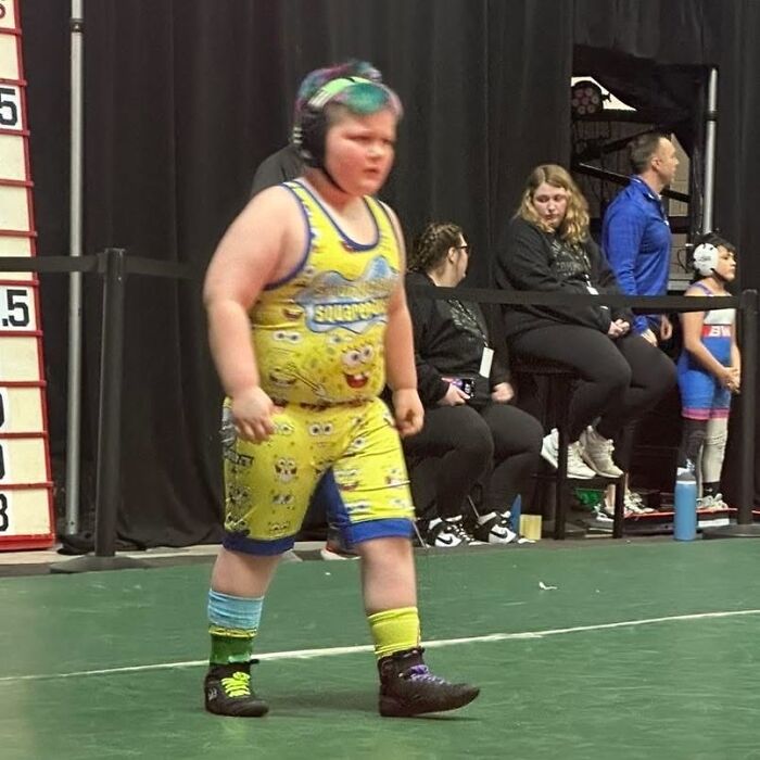 Child wrestler in mismatched socks and SpongeBob outfit preparing for a match in an unusual sports setting, a pic that makes no sense