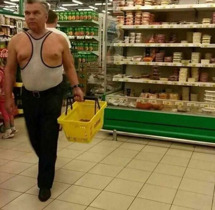 Man wearing an unusual sleeveless shirt walking in a grocery store aisle, holding a yellow shopping basket.