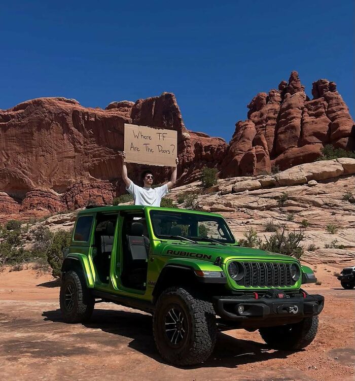 Dude With The Sign holding a cardboard message by a green off-road vehicle in a rocky desert landscape.